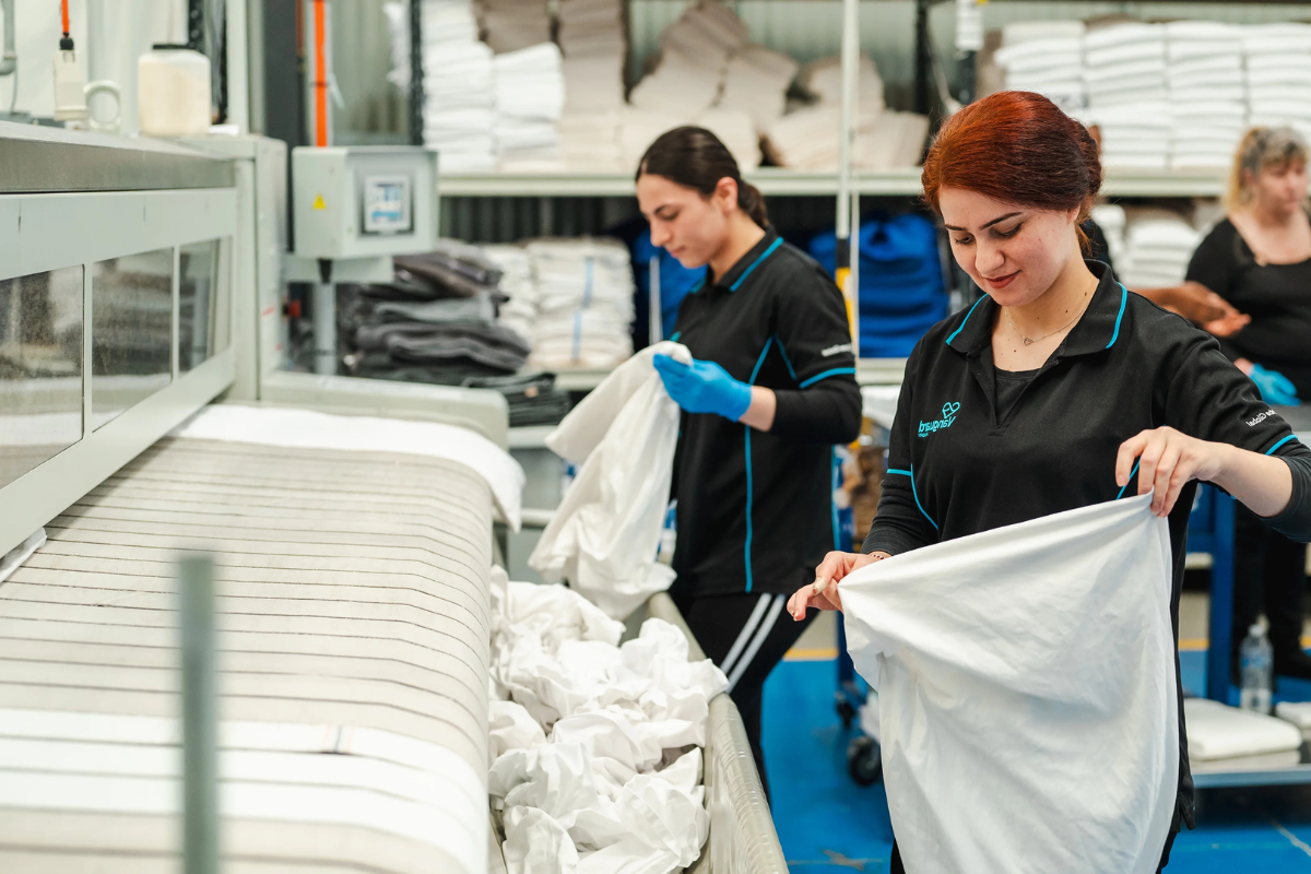 Workers in uniforms fold clean white linens at an industrial laundry facility with stacked shelves.